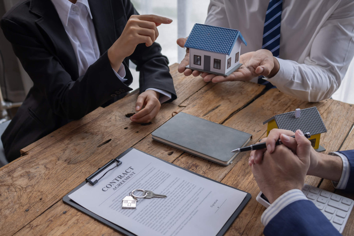 Sur une table en bois, trois personnes examinent une maquette de maison au toit bleu, des documents et une clé de maison tout en discutant de la gestion locative à Rennes
