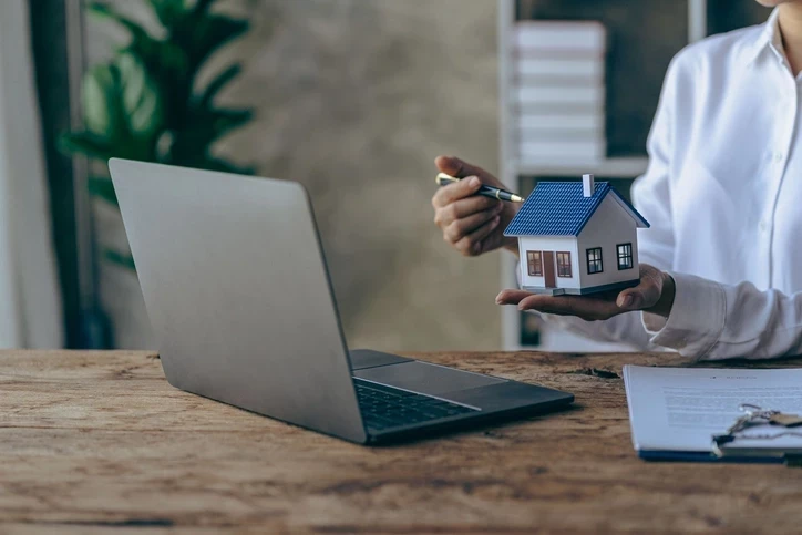 Une personne maintenant une maquette de bien immobilier, devant un ordinateur sur lequel est installé une gestion locative logiciel