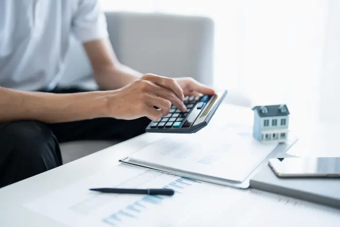 Personne utilisant une calculatrice sur des documents financiers avec une maquette de maison, illustrant l’analyse et la gestion d’un crédit immobilier.