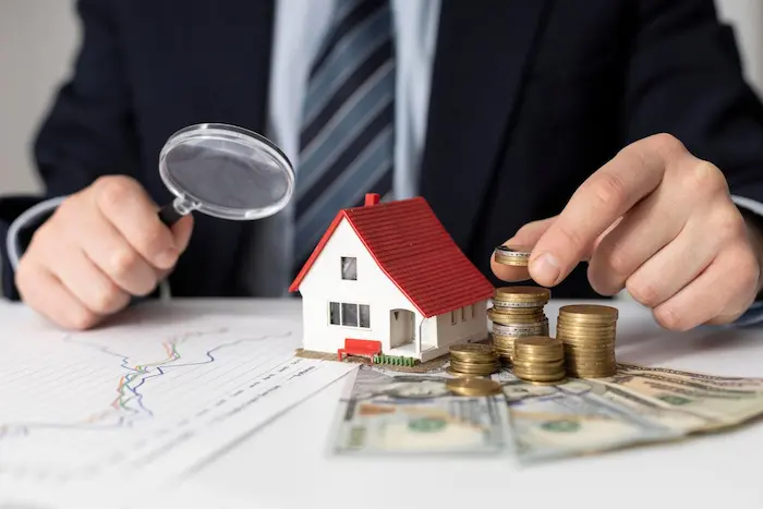 Un homme en costume examine une maisonnette avec une loupe et des piles de pièces pour expliquer comment financer investissement immobilier avec un prêt bancaire