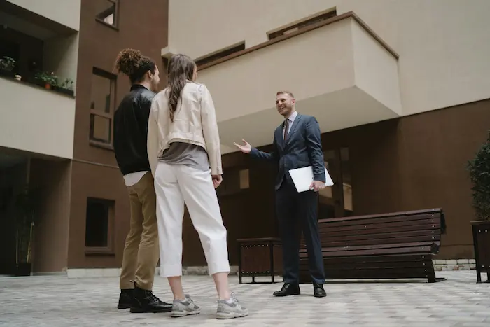 Courtier en costume présentant un dossier immobilier à des clients dans une cour moderne, image illustrant le rôle du courtier prêt immobilier 