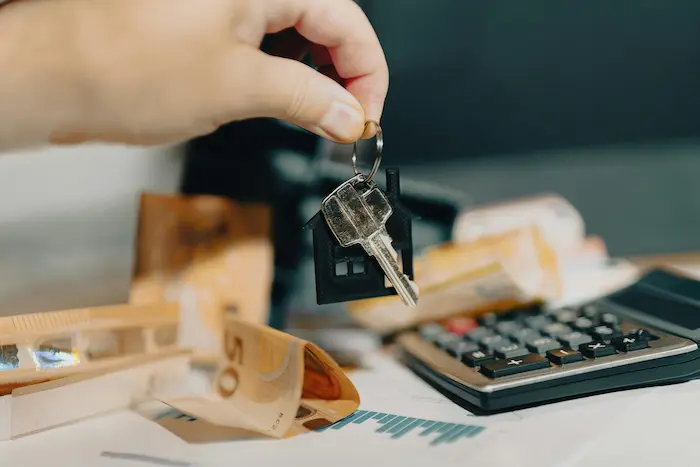 Main tenant une clé en forme de maison au-dessus de billets et calculatrice, image illustrant l’usage d’un simulateur d’encadrement des loyers.