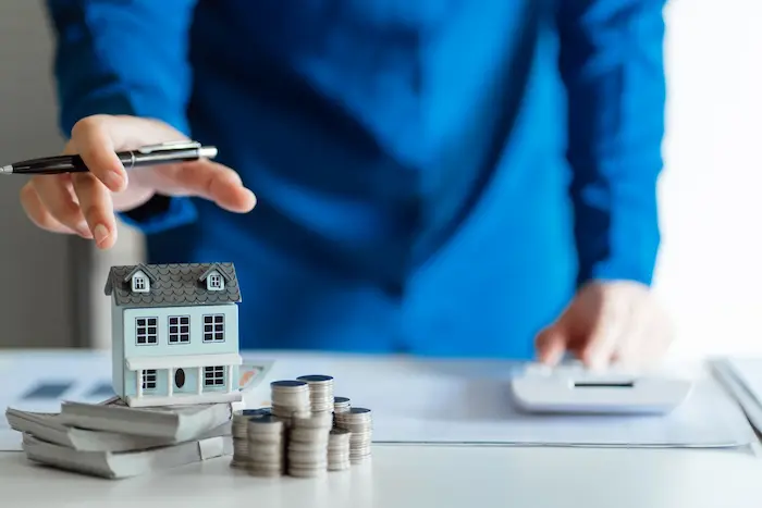 Personne en chemise bleue utilisant une calculatrice près d’une maquette de maison et piles de pièces, image illustrant le rachat de crédit immobilier.